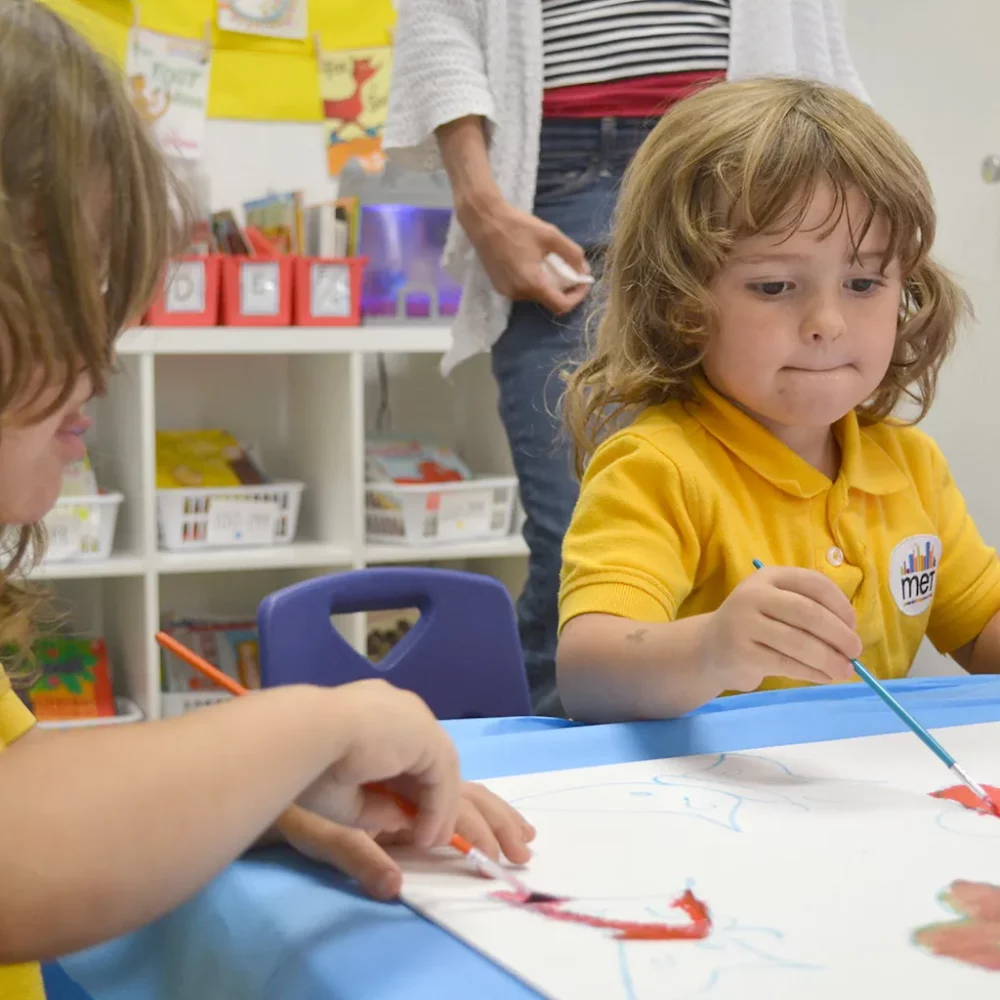 Small children sitting a table in their classroom painting with paintbrushes